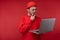 Photo of attractive man with beard in glasses and red clothing. Serious man holds laptop and wonders,  over red