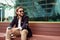 Photo of bearded brunet looking to side with glass in hand sitting on wooden bench against background of glass building