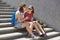 Photo of beautiful female teenagers sit on stairs outdoor, wears sunglasses and casual clothes, carry rucksacks, have rest after c