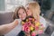 Photo of blonde mother and daughter sit couch hold flowers kiss smile make selfie indoors inside house apartment