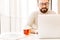 Photo of caucasian mature man 30s with beard and mustache drinking tea from glass, while working or browsing internet on laptop