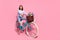 Photo of charming carefree young woman wear white sweater driving bike rising legs empty space smiling isolated pink