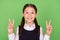 Photo of charming shiny school girl dressed formal clothes smiling showing v-signs  green color background