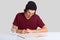 Photo of clever male college student rewrites information from book into notepad, prepares homework for tomorrow, being busy with