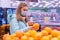 Photo of a cute girl in a medical mask and gloves choosing tangerines in the supermarket looking at camera. Shopping in