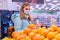 Photo of a cute girl in a medical mask and gloves choosing tangerines in the supermarket looking at camera. Shopping in