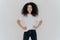 Photo of delighted curly woman keeps both hands on waist, smiles gently, has slim figure, wears white t shirt and black jeans,