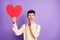 Photo of doubtful serious young guy wear yellow shirt arm chin holding big paper red heart isolated violet color