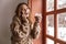 Photo of european woman 20s with brown hair talking on smartphone near window, and drinking glass of latte in cafe