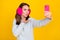 Photo of flirty happy young brunette woman wear ear warmer make selfie send air kiss isolated on yellow color background