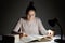 Photo of focused woman has hair knot, has serious clever expression in textbook, sits at desk with lamp, busy with exam preparatio