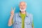 Photo of funky cute retired man wear vintage jeans waistcoat showing v-sign smiling isolated blue color background