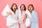 Photo of girlfriends 20s in home wear holding pillows and expressing excitement at slumber party, isolated over pink background