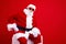 Photo of good mood funky grandfather in santa costume in glasses sit on white platform hand on waist isolated on red