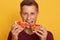 Photo of guy dressed in burgundy t shirt holding two pieces of pizza and eats fast food, enjoys delicious dish, male posing over