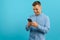 Photo of happy handsome man holding mobile phone in hand, texting. Young guy with big toothy smile posing on blue background