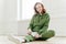 Photo of happy red haired woman in sweatsuit, white sneakers, takes rest after workout, sits on floor, stretches legs, has active