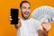 Photo of joyous man 30s in casual wear holding cell phone and fan of money, isolated over yellow background