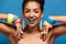 Photo of magnificent half-naked woman with colorful makeup smiling and demonstrating accessories on her arms, isolated over blue