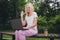 Photo of mature funny blond lady sit talk laptop wear white t-shirt trousers sit in park