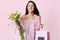 Photo of pleased brunette European lady holds gift bag and flowers, happy to recieve present on Womens Day, dressed in polka dot