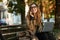 Photo of pleased girl using mobile phone and laptop while sitting on bench in sunlit alley