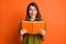 Photo portrait of surprised brunette girl keeping book laughing isolated on vivid orange color background