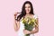 Photo of positive dark haired woman touches hair, smiles gently, has appealing look, holds first spring flowers, has red manicure