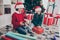 Photo of positive kids help santa sit floor prepare december presents wear x-mas hat jumper in decorated home indoors