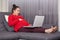 Photo of pregnant woman wears red sweater and maroon leggins keeps hand on tummy, opens mouth widely, lies on gray sofa in living