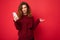 Photo of pretty asking upset young woman with curly hair wearing dark red sweater isolated on red background holding