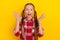 Photo of pretty impressed schoolgirl wear checkered shirt rising arms smiling isolated yellow color background