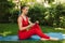 Photo of pretty woman in red jumpsuit sitting and using cellphone on yoga mat while practicing yoga in park