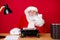 Photo of santa claus grey beard sit desk fist cheek vintage typewriter lamp paper book wear x-mas costume coat cap