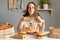 Photo of satisfied delighted woman wearing white T-shirt sitting at table in kitchen and eating pasta, posing with mouth full of