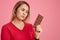 Photo of serious woman holds bar of cocoa chocolate, feels temptation, keeps to diet, dreessed in red outfit, isolated over pink