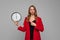 Photo of serious young woman pointing finger on big round clock holding in hand, she wonders how much time passed, standing in red