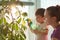 Photo of small kid holds spray bottle with water, waters green plant at home, helps his mother about house. Young housewife sprays