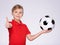 Photo of a smiling boy holding soccer ball and showing a big thumb up. posing at studio. Happy 8 years old kid in a red t-shirt