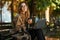 Photo of smiling woman working on laptop while sitting on bench in sunlit alley