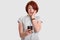 Photo of stupefied red haired woman in casual outfit holds mobile phone, recieves unexpected notification, concentrated in screen