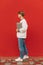 Photo of a stylish full-length guy standing on a background of a red wall with a laptop in his hands and wireless headphones and