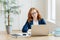 Photo of successful businesswoman with red hair, wears optical glasses for vision correction, sits at desktop in front of opened