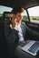 Photo of successful man wearing suit and earbud working on laptop, while back sitting in business class car