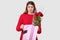 Photo of surprised terrified young woman has stupefied facial expression while opens gift bag, holds roses, dressed in red clothes