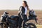 Photo of thoughtful Caucasian female likes riding motorbike, poses near her fast bike, has leather jacket on shoulder, keeps hand