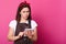 Photo of thoughtful girl with brown apron soiled with flour, holds gadget in hand and tries to find recipe for making cake with