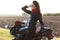 Photo of thoughtful young woman stands sideways near motorrbike, wears red bandana, sunglasses, leather jacket, looks pensively in