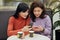 Photo of two busy curious good looking young females using their smartphones, chatting, sitting at table in cafeteria, drinking