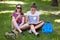 Photo of two female teenagers sit on green grass outdoor, read book and uses mobile phone for surfing social networks, wear sungla